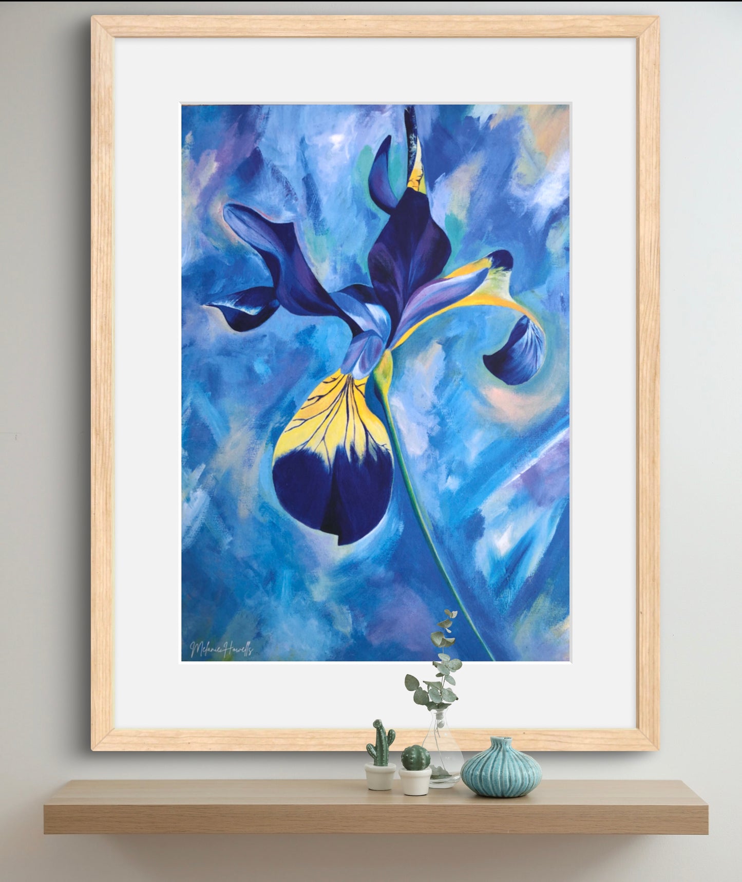 A framed abstract floral art print featuring a blue and yellow iris-like flower on a textured blue background, displayed on a shelf above small decorative plants and vases.