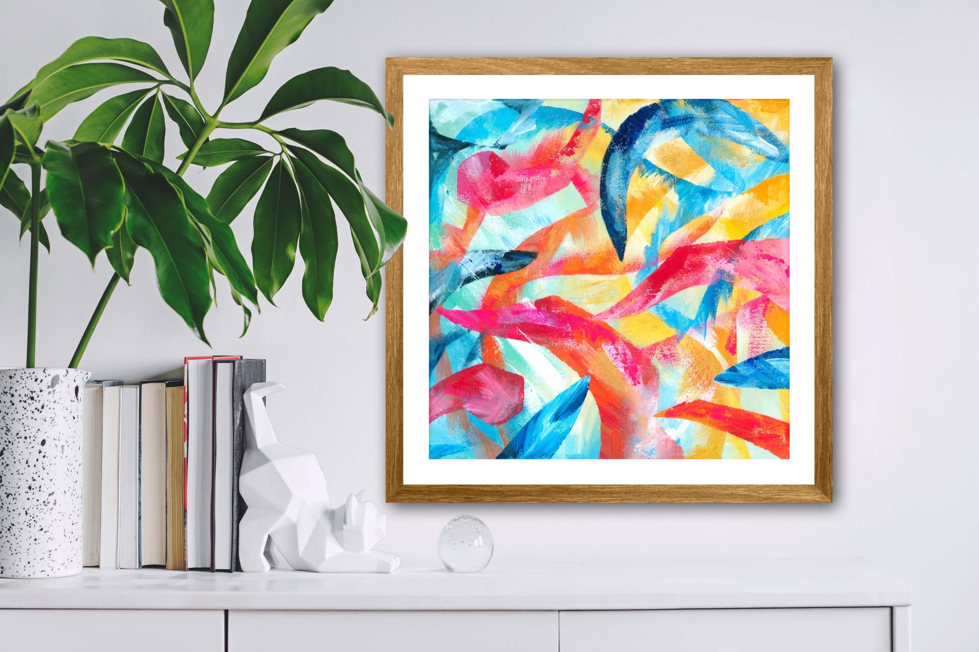 Adventure colourful abstract art print displayed in a natural wood frame on a white wall next to a shelf with a plant and books on it.