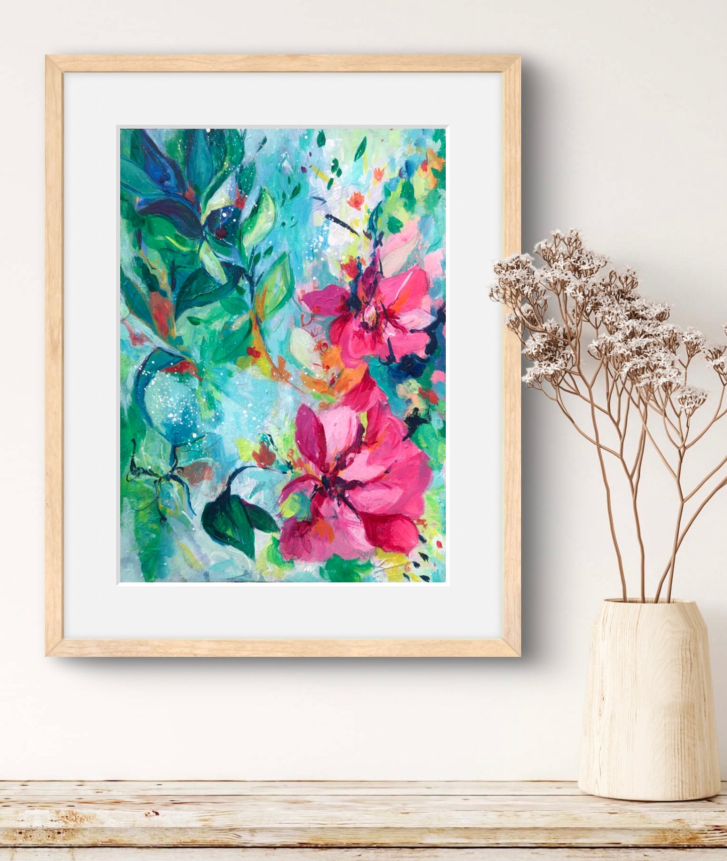 Framed colorful floral painting on a wall next to a vase with dried flowers.