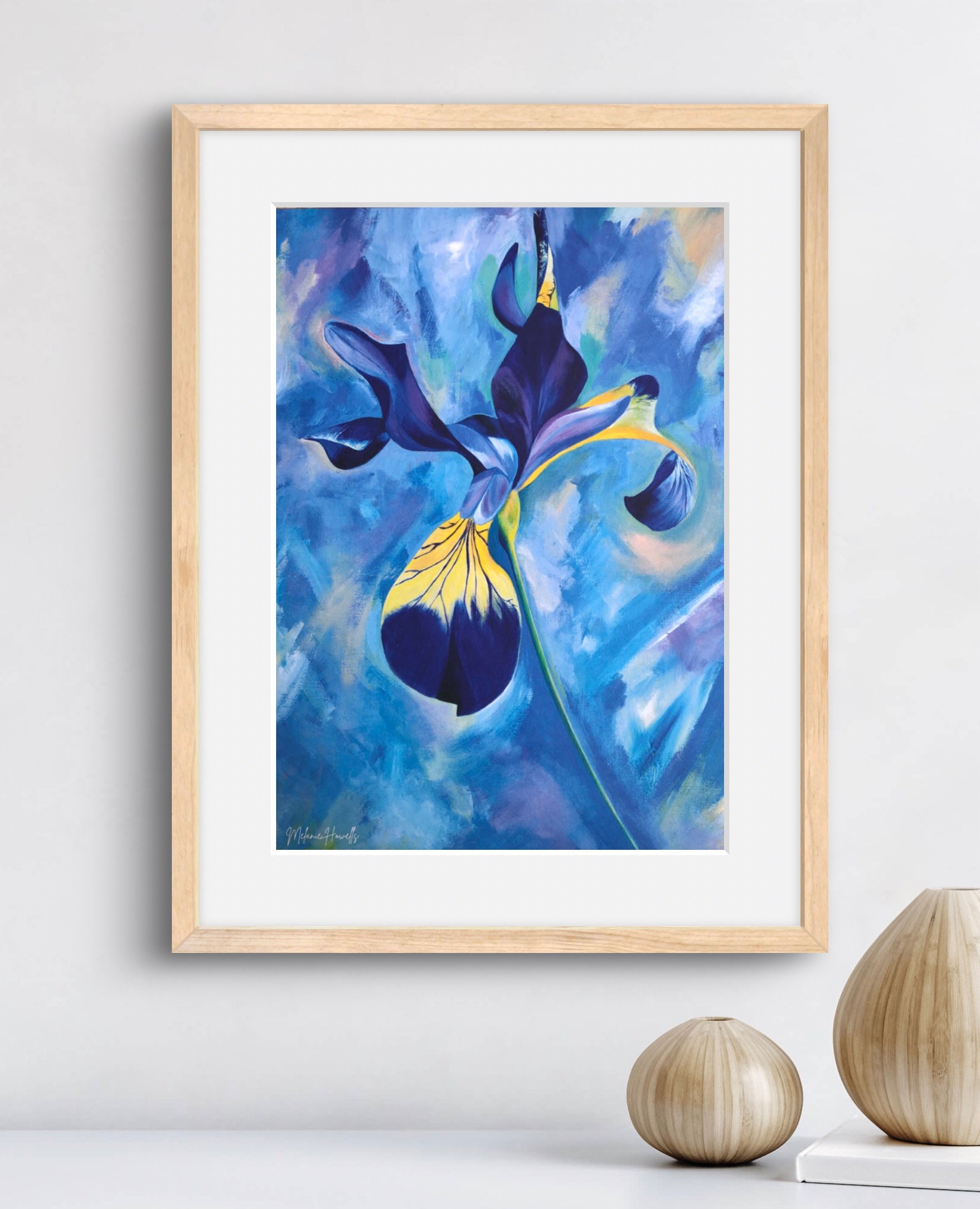Framed wall art featuring an expressive blue and purple iris painting with yellow accents, displayed in a light wood frame on a white wall above a surface with two decorative vases.