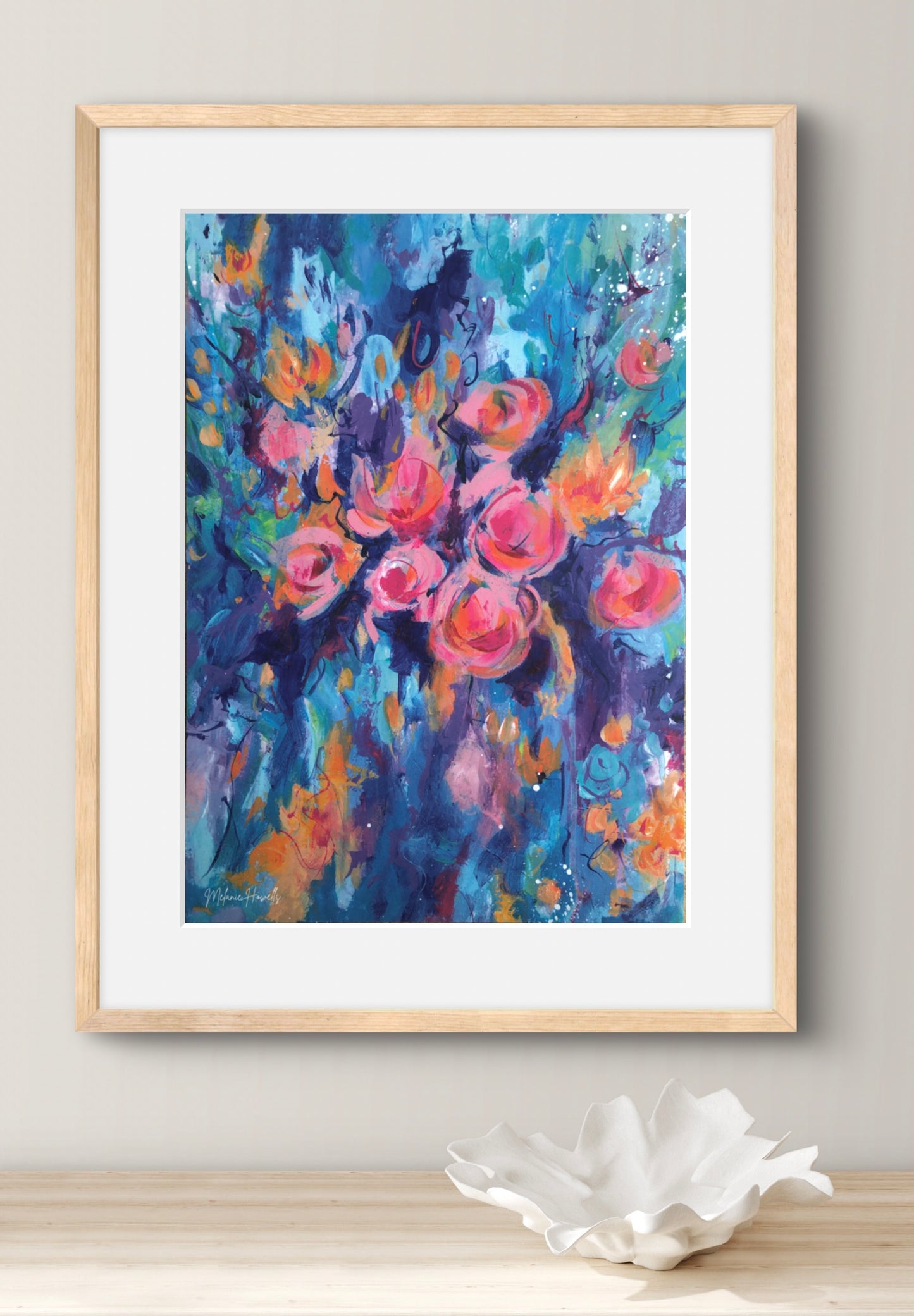 Framed floral wall art featuring an expressive painting of pink roses with blue, purple, and orange brushstrokes, displayed in a light wood frame with a white mat against a neutral wall above a white decorative object.