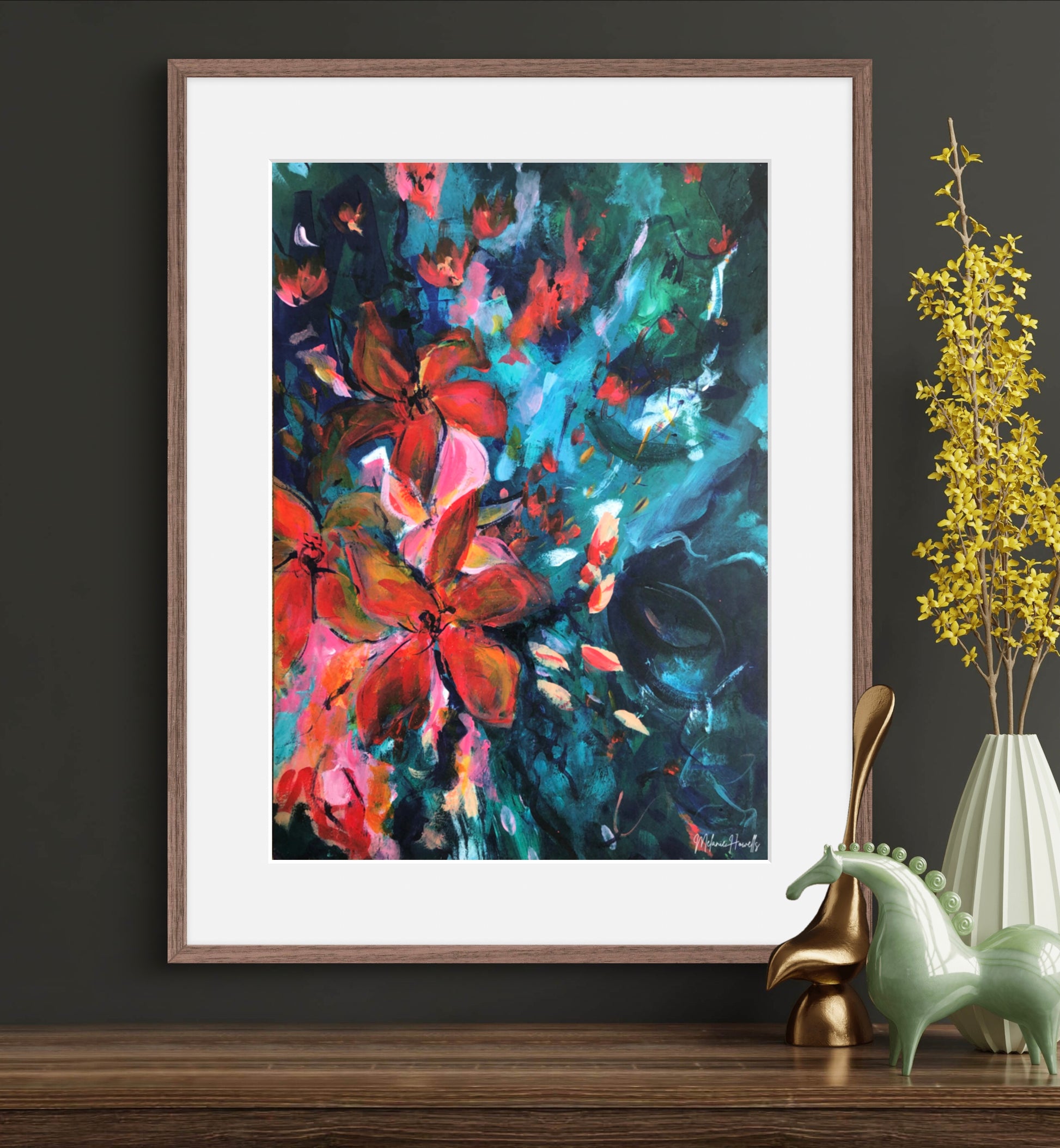 Framed abstract floral wall art hanging on a dark wall above a wooden surface, with decorative horse figurines and a vase of yellow stems beside it. The artwork features bold brushstrokes in red, pink, teal, blue, and black.