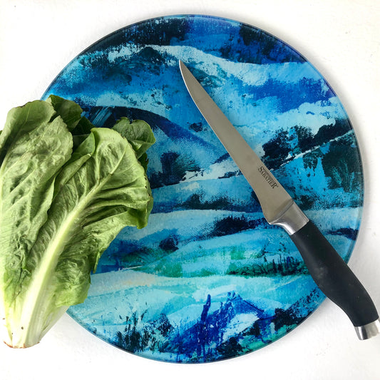 Round glass chopping board featuring a blue landscape design shown with a lettuce and knife