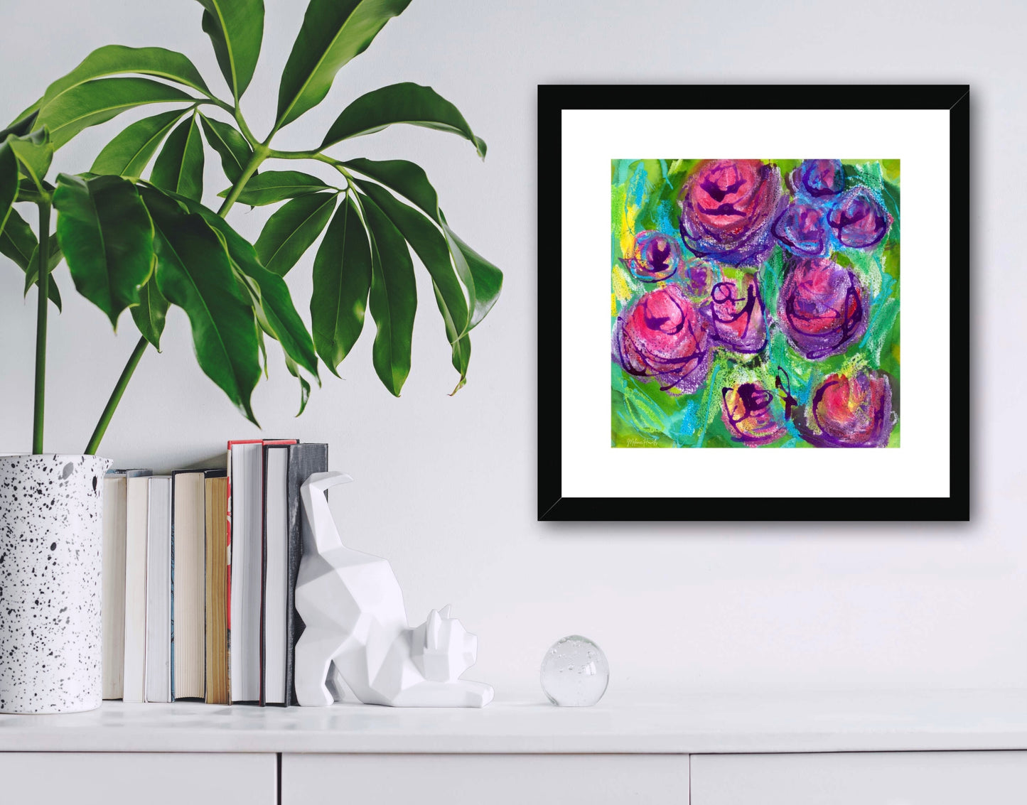 Rose Garden pink, purple and green vibrant floral giclee print shown framed in a black wooden frame above a white shelf with books and a plant..