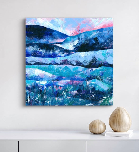An abstract landscape canvas print featuring layered blue and teal hills under a pink and light blue sky, displayed on a white wall above a white shelf with two decorative vases.