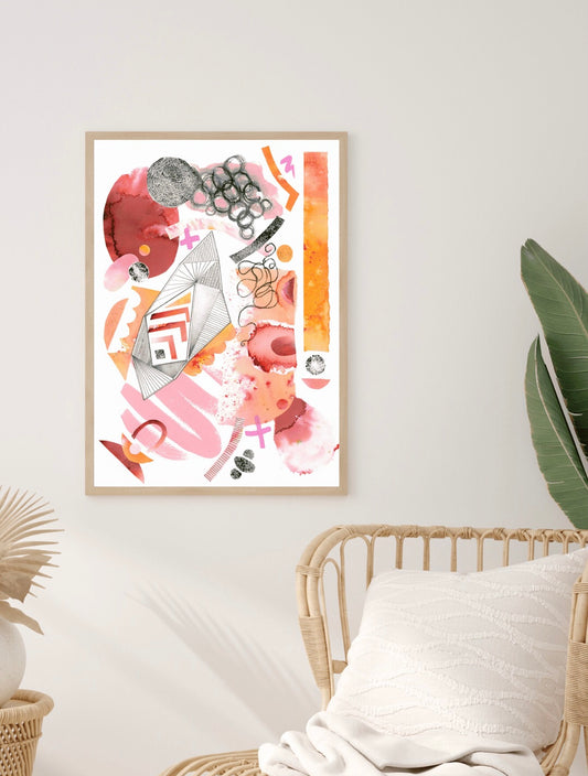 Catalyst pink and orange giclee wall art displayed in a natural wood frame on a cream wall next to a wicker chair