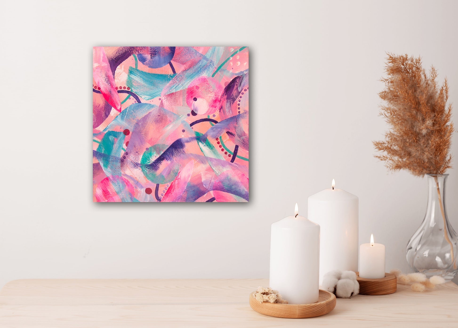 'Optimist' original acrylic painting in shades of pink measuring 30x30cm displayed on a white wall next to a vase and candles.