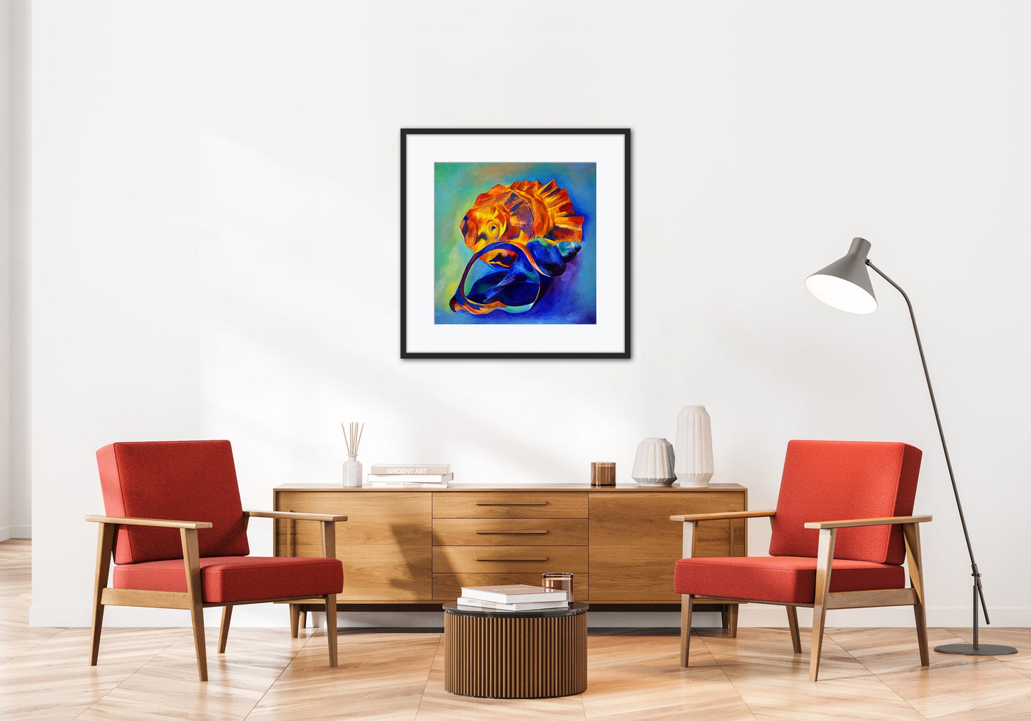 The Beachcomber colourful seashell art print displayed in a black frame on a white wall in a contemporary lounge with 2 red chairs and a wooden sideboard.